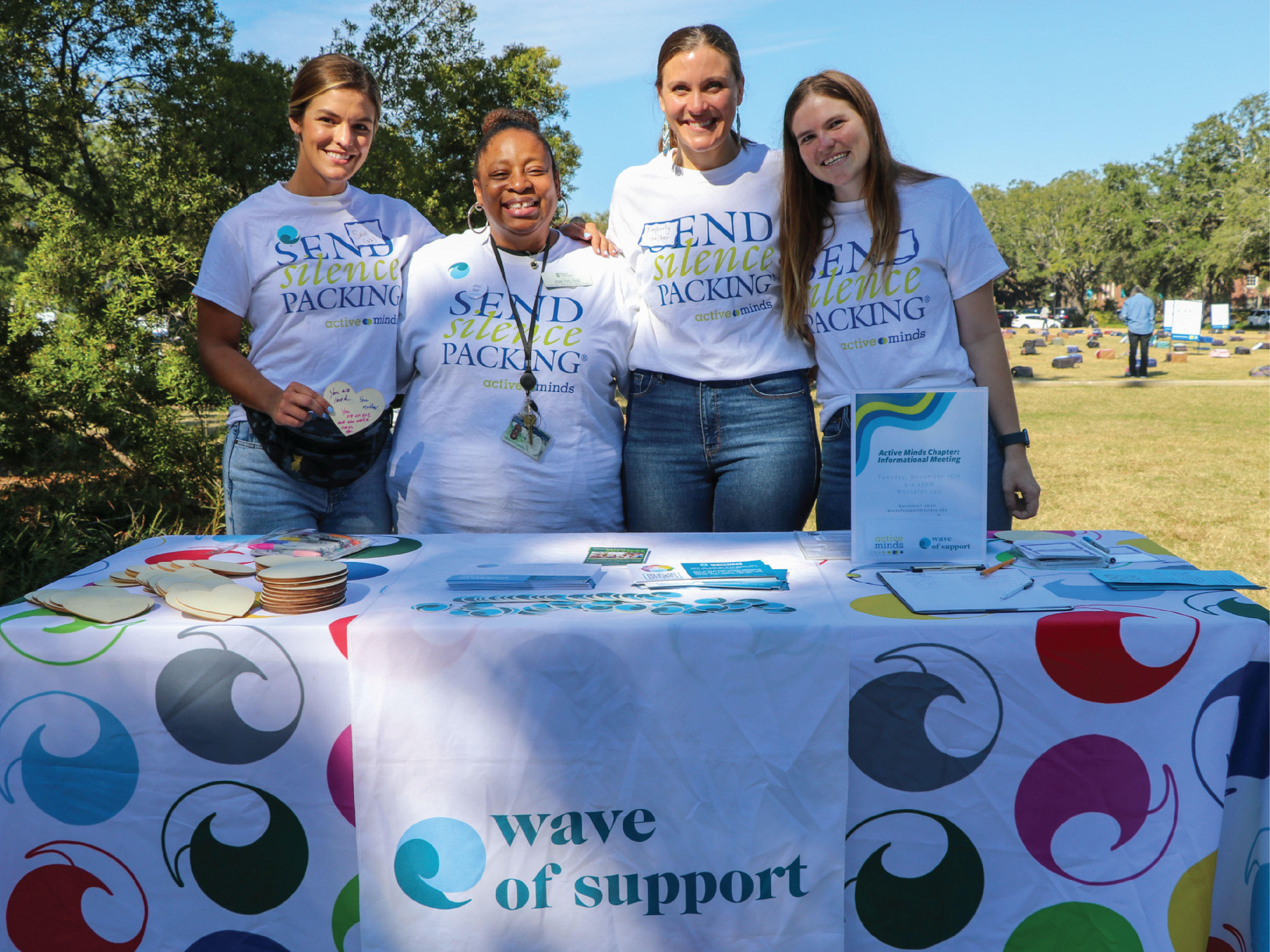four staff members smile, standing at the Wave of Support table during the Send Silence Packing event